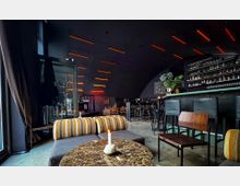 Moderne Bar mit dunkel gehaltenem Interieur, beleuchtet durch rote, hängende Lichtleisten. Im Vordergrund ein Marmortisch mit Kerze, dahinter Polsterbänke und Stühle; im Hintergrund eine lange Theke mit Barhockern und einer Regalwand mit Flaschen.