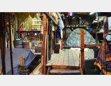 Innenraum einer rustikalen Werkstatt oder Scheune mit Holzwänden und -boden. Im Vordergrund sichtbar ist ein kleines Podest mit einem Tarnzelt und umliegenden Gegenständen wie Werkzeug, Regale, Fässer und aufgehängte Kleidungsstücke.