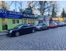 Straßenansicht mit mehreren kleinen Gewerbebauten, darunter Schilder- und Versicherungsdienste, mit bunten Fassaden und Reklameschildern. Davor stehen Autos auf einer gepflasterten Straße geparkt, und im Hintergrund sind Bäume sichtbar.