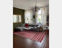 Wohnzimmer mit einem grauen Ecksofa, einer roten gemusterten Teppichbrücke und einem kleinen Couchtisch. Die Wände sind teilweise olivgrün gestrichen, mit einem großen Spiegel und Kunstwerken dekoriert; große Fenster mit weißen Vorhängen lassen Tageslicht herein.