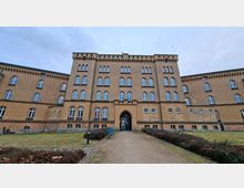 Frontansicht eines großen, mehrstöckigen Backsteingebäudes mit vielen symmetrisch angeordneten Rundbogenfenstern und einem mittigen Spitzbogen-Eingang. Ein gerader Kiesweg führt durch eine Rasenfläche mit niedrigen Hecken zur Eingangstür unter bewölktem Himmel.