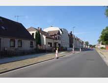 Straßenansicht eines Wohngebiets mit einer Reihe von Häusern auf der linken Seite, teils aus älteren Ziegel- und Putzfassaden. Auf dem Bürgersteig und der Fahrbahn stehen rot-weiße Absperrungen. Der Himmel ist klar und blau, und an der rechten Straßenseite befinden sich Rasenflächen und vereinzelte Bäume.