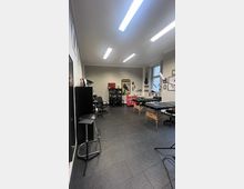 Ein heller Tattoostudio-Raum mit grauem Boden, schwarzen Möbeln und mehreren Behandlungsliegen. An den Wänden hängen Dekorationen wie Bilder, eine Gitarre und Poster, während der Raum durch helle Deckenbeleuchtung ausgeleuchtet wird.