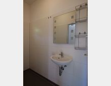 Kleines Badezimmer mit einem weißen Waschbecken, einem rechteckigen Spiegel und weißen Wandfliesen. Rechts neben dem Waschbecken ist ein Metallregal an der Wand angebracht.