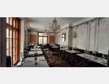 Eleganter Restaurant-Speisesaal mit hohen Holzfenstern, marmorierten grauen Wandpaneelen und dunkler Sitzbank entlang der Wand. Mehrere gedeckte Tische mit schwarzen Stühlen stehen auf Fischgrätparkett, darüber hängen verzierte Kronleuchter und Wandlampen.