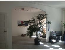 Wohnzimmer mit weiß gestrichenen Wänden, einem großen Zimmerbogen, einer Zimmerpflanze in einem schwarzen Topf und einem schwarzen TV-Regal mit Büchern und CDs. Im Hintergrund ist ein Esstisch in einem angrenzenden Raum zu sehen.