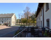 Ein gepflasterter Hof mit zwei Gebäuden, eines aus hellen Putzfassaden und eines mit roten Backsteinakzenten. Im Hintergrund stehen Bäume in einer parkähnlichen Umgebung, während im Vordergrund eine Terrasse mit Sitzmöbeln und Pflanzen zu sehen ist.