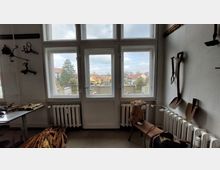 Ein Raum mit einer großen Fensterfront, die auf eine Terrasse und dahinterliegende bunte Häuser blicken lässt. An der Wand hängen landwirtschaftliche Werkzeuge wie Schaufeln, während ein Holzstuhl mit geflochtener Tasche und ein Tisch mit Holzstücken den Raum ergänzen.