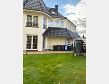 Ein modern gestaltetes zweistöckiges Wohnhaus mit cremefarbener Putzfassade und einem dunklen Ziegeldach. Der Vordergrund zeigt einen kleinen Garten mit Rasen, mehreren Mülltonnen nahe einer gepflasterten Terrasse und einem angrenzenden Gitterkäfig.