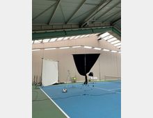 Innenansicht einer Tennishalle mit blauem Bodenbelag. Im Vordergrund befinden sich Fotoausrüstungen wie ein schwarzer Vorhang auf einem Stativ, eine Leiter und ein weißer Hintergrund, während das Dach Stahlträger und Oberlichter zeigt.