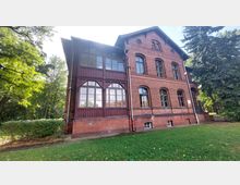 Backsteingebäude mit großen, dunkelroten Holzelementen und Fenstern an der linken Fassade, umgeben von Rasen und Bäumen. Das Gebäude hat zwei Stockwerke und einen sichtbaren Dachgiebel.