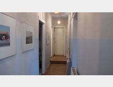 Ein schmaler, heller Flur in einem Wohngebäude mit Holzboden und einfachen weißen Wänden. An den Wänden hängen gerahmte Bilder, und am Ende des Flurs führt eine weiße Tür zu einem angrenzenden Raum.