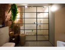 Badezimmer mit Backsteinwand, gemusterten Fliesenboden und verglaster Dusche mit schwarzem Metallrahmen; links hängen Pflanzen, daneben stehen geflochtene Körbe.