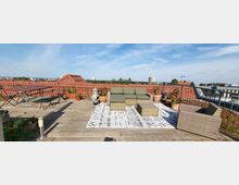 Dachterrasse mit Holzboden und einem Sitzbereich aus Rattanmöbeln auf einem gemusterten Teppich, umgeben von Pflanzen in Töpfen; im Hintergrund sind rote Ziegeldächer und hohe Gebäude zu sehen, unter blauem Himmel.