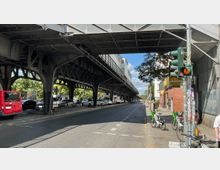 Straßenansicht mit einer erhöhten metallenen U-Bahn-Brücke, die über die Fahrbahn verläuft. Unter der Brücke sind parkende Autos und Fahrradbügel zu sehen, rechts befindet sich ein Bürgersteig mit einem Ampelmast sowie Leihfahrrädern. Im Hintergrund stehen Bäume und Gebäude.