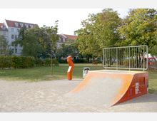 Skatepark-Bereich in einer grünen Parklandschaft mit Bäumen und Rasenflächen, umfasst eine orangefarbene Rampe mit Geländer und eine auffällige orange Skulptur. Im Hintergrund sind Wohngebäude mit roten Dächern sichtbar.