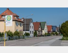 Eine ruhige Wohnstraße mit Einfamilienhäusern, die Satteldächer in unterschiedlichen Farben haben, einige davon mit Solarpanelen. Im Vordergrund steht ein gelber Bushaltestellenschild mit der Aufschrift "Rathenow Fortschritt".