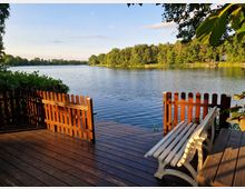 Holzterrasse mit einem weißen Gartenstuhl und einem niedrigen Holzzaun, direkt am Ufer eines ruhigen Sees gelegen, umgeben von üppigem Grün und Bäumen im Hintergrund. Die Abendsonne beleuchtet die Szene, und der Himmel ist leicht bewölkt.
