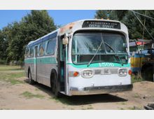 Ein älterer silberner und grüner Bus mit der Aufschrift „GMC“ und der Nummer „4506“ steht auf einer grasbewachsenen, teils sandigen Fläche, umgeben von Bäumen und weiteren Fahrzeugen im Hintergrund. Die Frontscheibe zeigt die Beschriftung „Special Service“.