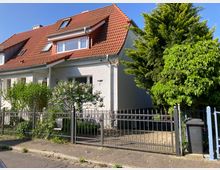 Einfamilienhaus mit weißer Fassade und rotem Satteldach, Teil einer Doppelhaushälfte. Vor dem Haus befindet sich ein Vorgarten mit blühenden Sträuchern und ein schwarzer Metallzaun, flankiert von hohen Bäumen.