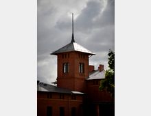 Backsteingebäude mit einem Turm und Spitzdach, verziert mit einem schmalen Mast. Der Himmel ist bewölkt, und im Vordergrund ist ein Teil eines Baums sichtbar.