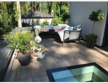 Moderne Dachterrasse mit Holzdeck, schwarzen Loungemöbeln und weißen Kissen sowie mehreren Pflanzkübeln und hängenden Laternen. Im Hintergrund sind ein flaches Nebengebäude und dichter Wald zu sehen; am Terrassenrand befindet sich ein verglaster Bodenausschnitt.