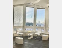 Bright modern office lounge with curved floor-to-ceiling corner windows fitted with blinds, overlooking a city skyline with a tall TV tower. Two white swivel chairs and small curved side tables sit on dark wood flooring beside low radiators under the windows.