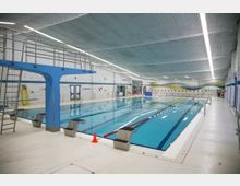 Hallenbad mit Schwimmbecken, Sprungturm und Startblöcken; geflieste Wände, rutschfester Bodenbelag und Deckennetz zum Schutz. Im Hintergrund sind Türen, Schwimmhilfen und Wandgrafiken sichtbar.