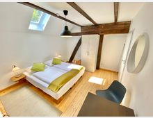 Helles Schlafzimmer mit Holzboden, einem Doppelbett mit weißer Bettwäsche und grünen Kissen, schräger Decke mit sichtbaren Holzbalken, einer Dachluke, einem weißen Schrank und einem kleinen Schreibtisch mit Stuhl.