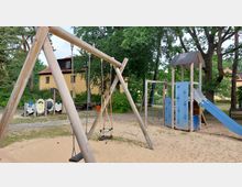 Spielplatz mit Holzschaukel, Klettergerüst mit blauen Elementen und Rutsche auf einem Sandboden. Im Hintergrund sind Bäume, Büsche und ein gelbes Gebäude mit roten Dachziegeln zu sehen.