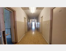 Ein einfacher, langer Flur mit beige-weißen Wänden, Türen auf beiden Seiten und einem Boden aus hellem Laminat. Am Ende des Flurs befindet sich ein Fenster, das Tageslicht hereinlässt.