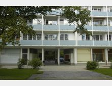 Mehrstöckiges Wohngebäude mit blauen Balkonen und weißen Fassaden, im Erdgeschoss ein Durchgang zu einem Hinterhof sowie Schaufenster von Geschäftsräumen. Im Vordergrund befinden sich grüne Rasenflächen und Bäume.