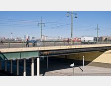Straßenbrücke mit Gehweg und Laternen, darunter eine gelbe Betonunterführung und Pfeiler. Im Hintergrund sind moderne Gebäude und der Berliner Fernsehturm sichtbar, bei klarem Himmel.