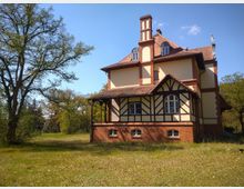 Fachwerkhaus mit cremefarbenen Wänden und dunklen Holzbalken, kombiniert mit roten Backsteinelementen und einem Ziegeldach. Das Haus steht auf einer grünen Wiese, umgeben von Bäumen, unter klarem, blauem Himmel.