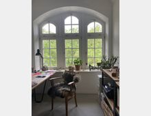 Ein heller Arbeitsbereich mit einem großen, dreiteiligen Rundbogenfenster, das auf grüne Bäume blickt. Der Raum enthält einen Schreibtisch mit Arbeitsmaterialien, einen Holzstuhl mit Fellauflage, Zimmerpflanzen auf der Fensterbank und ein Regal voller Bücher und Gegenstände.