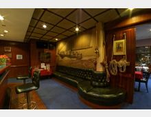 Innenraum einer maritim gestalteten Bar mit Holzwänden, blauer Teppichboden und einer langen, gepolsterten Ledersitzbank. An der Wand hängt ein großformatiges Wandbild eines Segelschiffes, daneben Seile, eine Messingglocke und maritimes Dekor.