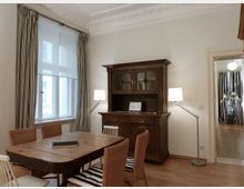 Esszimmer in einem Altbau mit hohen Decken und Stuckverzierungen. Zu sehen sind ein Holztisch mit gemischten Stühlen, ein antiker Holzschrank, zwei Stehlampen, ein Fenster mit gestreiften Vorhängen und ein Durchgang zu einem weiteren Raum.