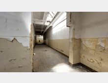 Ein leerer, verfallener Korridor mit abblätternder Farbe an den Wänden und einem Boden aus verwitterten Dielen. Die Decke zeigt freiliegende Rohre und eine Lichtöffnung, durch die Tageslicht einfällt.