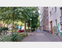 Straßenszene in einem Wohnviertel mit Bäumen, grauen Wohnhäusern und einem Gehweg mit Kopfsteinpflaster. Im Hintergrund ist ein gelber Doppeldeckerbus auf der Straße sowie geparkte Autos zu sehen; einige Hauswände sind mit Graffiti versehen.