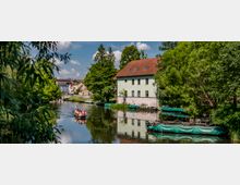 Eine Flusslandschaft mit einem traditionellen hölzernen Ausflugsboot, in dem Menschen sitzen. Am Ufer stehen ein großes hellgrünes Gebäude mit rotem Dach und mehrere Bäume, daneben liegen Boote mit grünen Abdeckungen. Im Hintergrund sind bunte Häuser einer Stadt zu sehen.