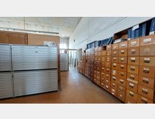 Ein Raum mit einer langen Wand aus Holzschubladen und Aktenordnern obenauf, gegenüber einer Reihe metallener Flachablageschränke. Im Hintergrund führt ein schmaler Gang zu großen Fenstern, die Tageslicht hereinlassen.