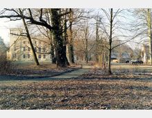 Parklandschaft mit kahlen Bäumen im Winter, einem schmalen Weg, der durch das Gelände führt, und einem historischen hellen Gebäude im Hintergrund. Rechts sind geparkte Autos und ein weiteres Gebäude zu sehen.