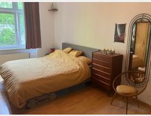 Schlafzimmer mit einem Bett mit senfgelber Bettwäsche, einer Holzkommode, einem runden Holzstuhl und einem hohen Spiegel mit gebogenem Rahmen. Ein Fenster mit braunem Vorhang und ein kleines Wandregal sind an der linken Wand zu sehen.