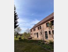 Historisches Backsteingebäude mit mehreren schmalen, bogenförmigen Fenstern und einem roten Ziegeldach, umgeben von einem gepflegten Garten mit Blumenbeeten und Rasenflächen. Im Hintergrund sind Bäume und eine offene Landschaft unter einem klaren blauen Himmel sichtbar.
