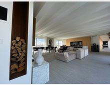 Modernes Wohnzimmer mit offener Raumaufteilung, großen Fenstern und hellen Wänden. Im Vordergrund eine eingelassene Holzablage, eine graue Sofalandschaft und ein Marmorsockel; im Hintergrund eine Küche mit Holzelementen und ein Tisch mit Stühlen.