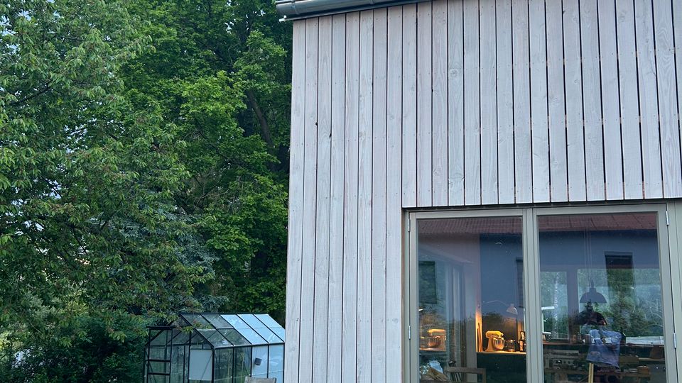 Außenansicht eines hellen Holzhauses mit steilem Metalldach und großem Fenster zur Gartenseite. Daneben liegt eine Wiese mit Sitzmöbeln, im Hintergrund stehen hohe Bäume und ein kleines Gewächshaus.