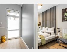 Blick in ein modernes Schlafzimmer mit Doppelbett, hoher gepolsterter Wandverkleidung als Kopfteil und heller Holzoptik am Boden. Links führt ein schmaler Flur zu einer Glastür, rechts stehen ein kleiner runder Beistelltisch mit Lampe und ein Heizkörper an der Wand.