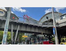 Bahnhofsanlage in Berlin mit Hochbahnbrücke aus Stahl, darunter eine Straßenszene mit Ampeln, Verkehrsschildern und Fahrzeugen. Im Hintergrund ein halbrundes Glasdach und ein blauer Himmel mit wenigen Wolken.