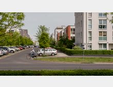Wohngebiet mit mehrstöckigen, modernen Wohngebäuden und parkenden Autos entlang der Straße. Im Vordergrund ein Radfahrer auf einer asphaltierten Straße, umgeben von gepflegten Grünflächen und Bäumen.