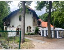 Ein zweistöckiges Gebäude im traditionellen Stil mit weißer Fassade, Fachwerkelementen und rotem Ziegeldach, umgeben von Bäumen. Im Vordergrund befinden sich ein grüner Zaun, ein Hinweisschild, ein gepflasterter Weg und ein angrenzendes weiß-grau gestreiftes Festzelt.